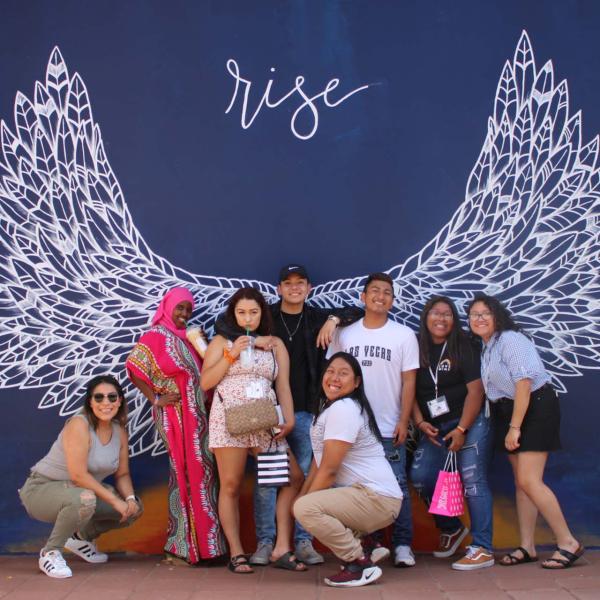 A group of students standing in front of a mural with dark blue background and white angel wings painted with the word "Rise" above them