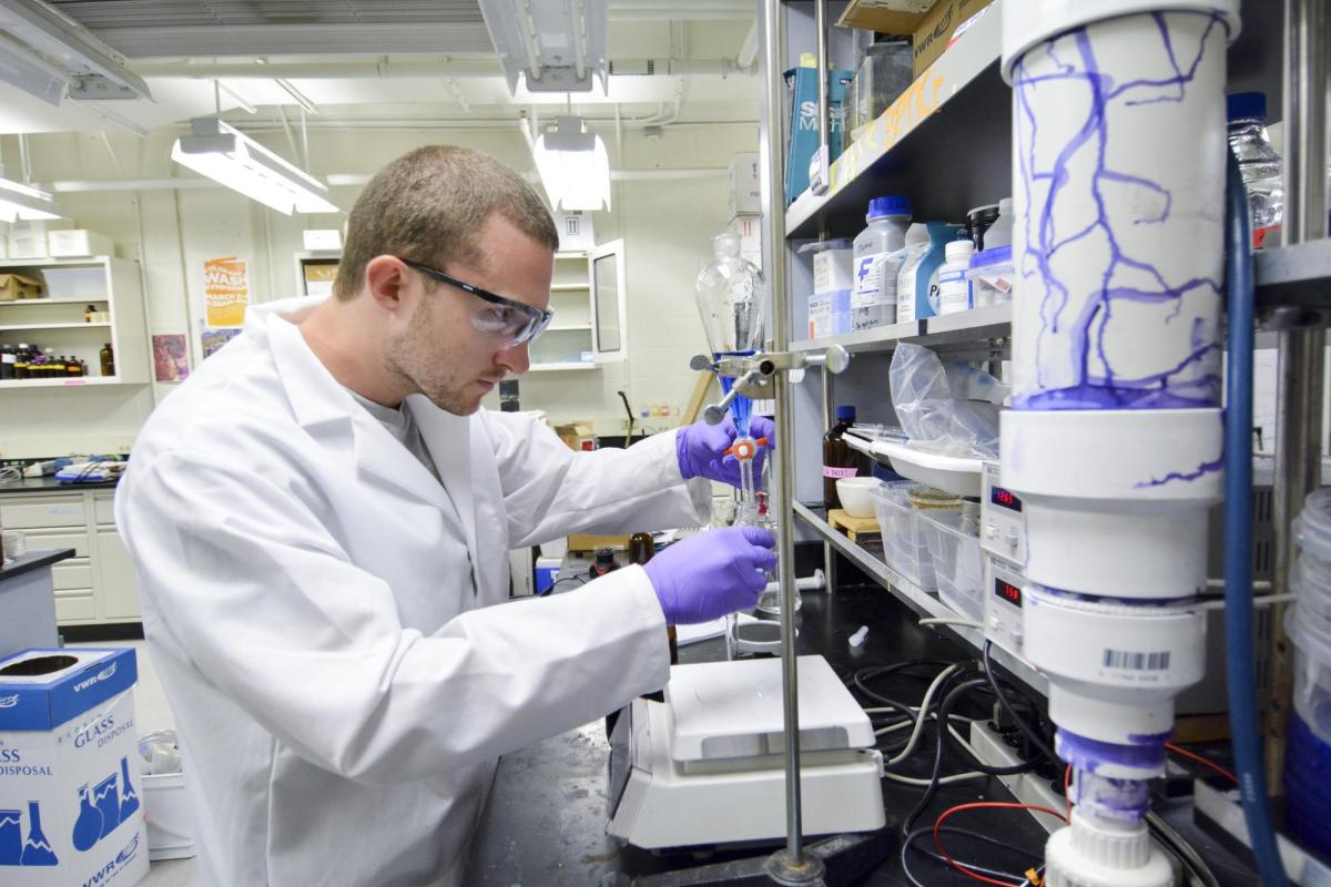 A student works with technical chemistry equipment in full lab regalia.