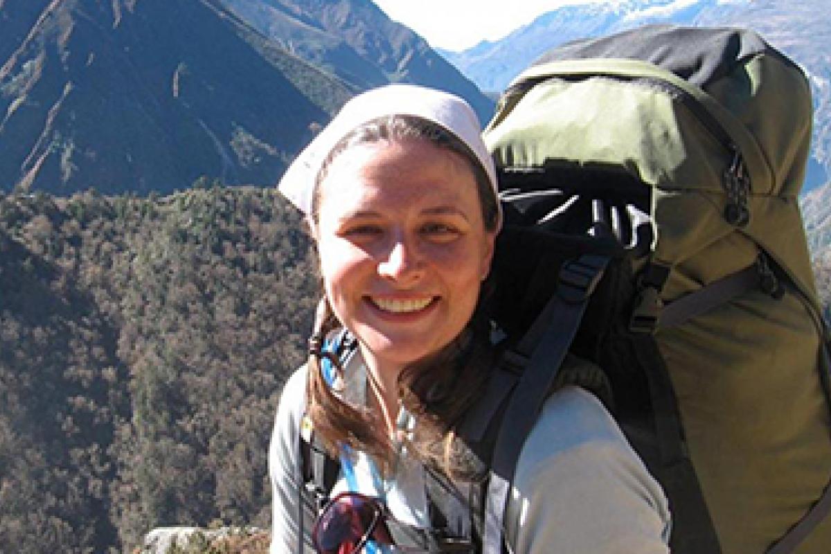 A student wearing a backpack smiles at the camera with wilderness behind her.