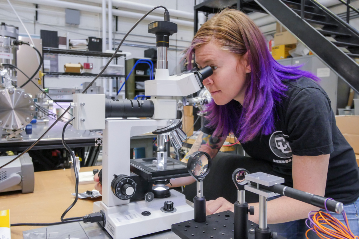 A physics student stares into a microscope, which is situated in a cluttered lab.