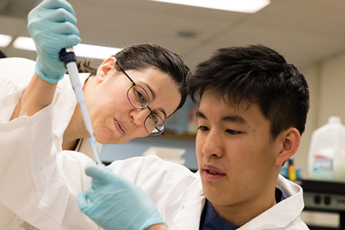 A student in lab dress uses a dropper to place a liquid in another contained while a faculty member watches.