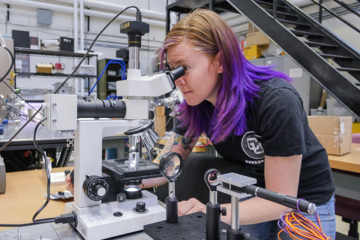 A student stares into a microscope while considerable physics lab equipment surrounds.