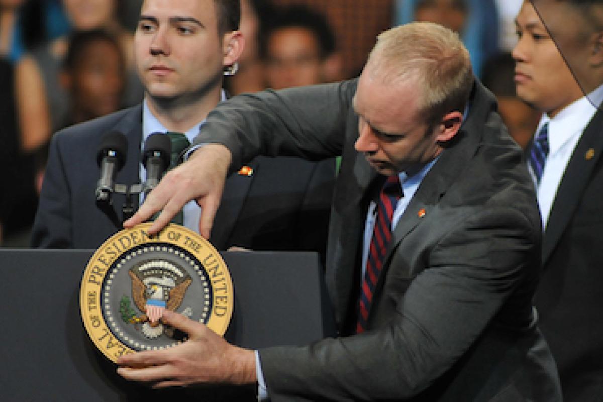 A man adjusts a symbol that says "President of the United States" on a podium while two other men in secret service garb look on.