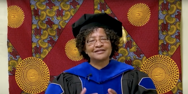 Donna Auguste giving a virtual commencement speech, dressed in graduation regalia.