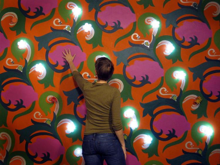 Woman reaching toward brightly colored painted wall with white flowers with lights.