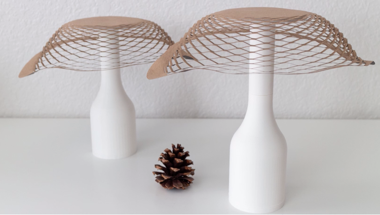 Two Sensing Kirigami lampshades sit on a table with a pine cone between them.