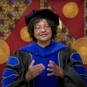 Donna Auguste giving a virtual commencement speech, dressed in graduation regalia.