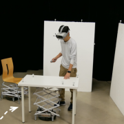 Person with VR goggles touches a table that has a Roomba with a mechanical scissor lift below. 