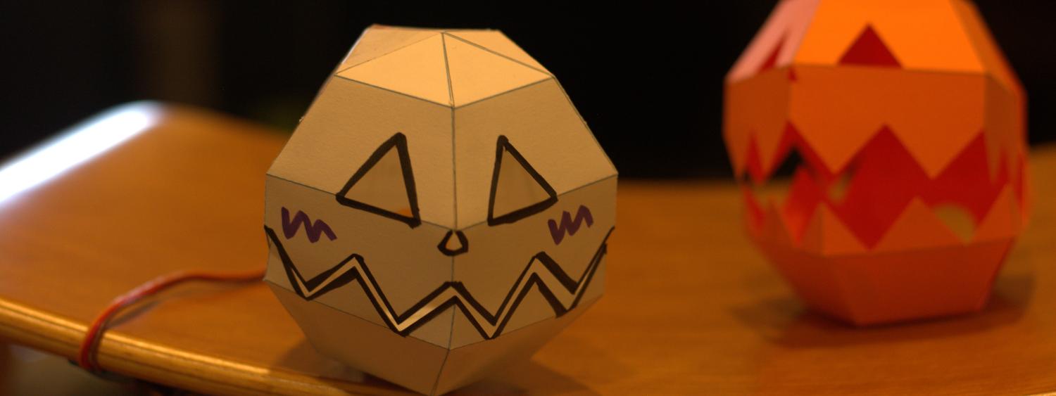 Two paper robots shaped like Jacko lanterns sit on a table. One is white and one is orange.