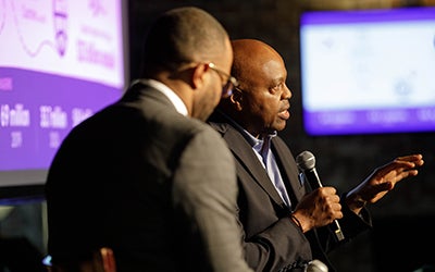 Two men in suits speak onstage at the event.