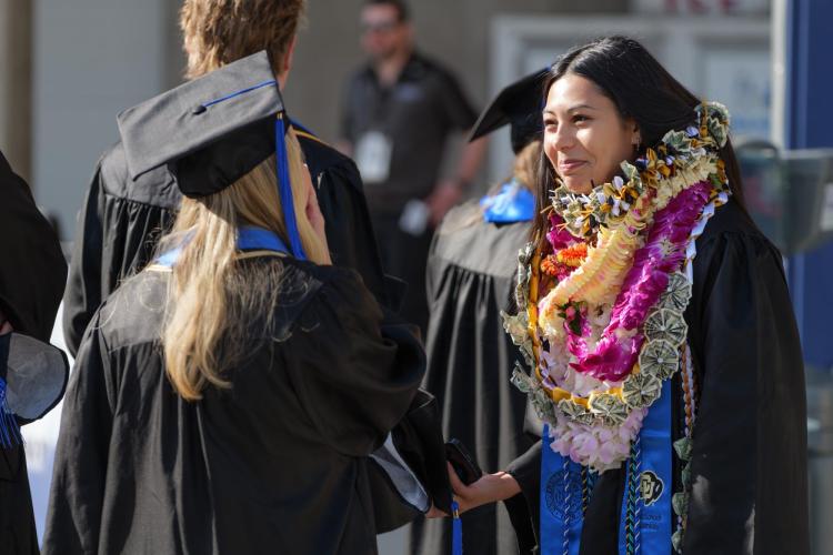 A student wearing a collection of leis and her regalia smiles while in conversation with another graduate. 
