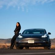 Assistant Professor Kyri Baker in the early morning light leaning against her Tesla, which is parked between two windmills.