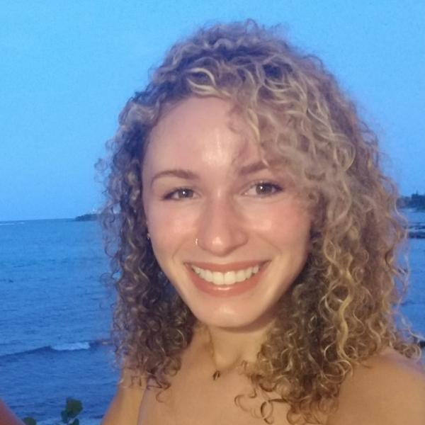 A picture of a person with blonde curly shoulder-length hair smiling at the camera standing outside