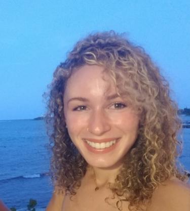A picture of a person with blonde curly shoulder-length hair smiling at the camera standing outside