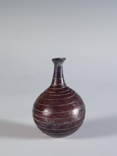 Photograph of a reddish glass vessel with a round body and base with a relatively short and narrow neck and outward-tapering mouth, from the side against a neutral gray background.