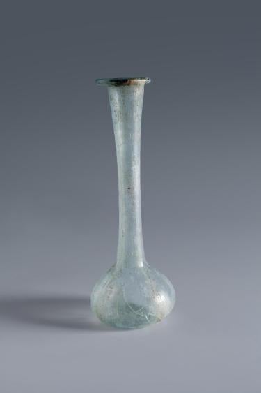 Photograph of a clear glass vessel with a squat globular body and a tall, narrow neck that flares toward the top to a rounded mouth, from the side against a neutral gray background.