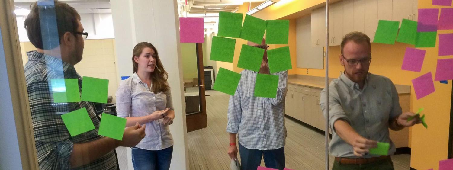 Three men and one woman put sticky notes on a glass wall.