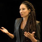 Ruha Benjamin, in professional attire, speaking before a black backdrop.