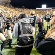 A group of CMCI students shoots video and photos amid a group of players on the sidelines of the stadium during a football game.