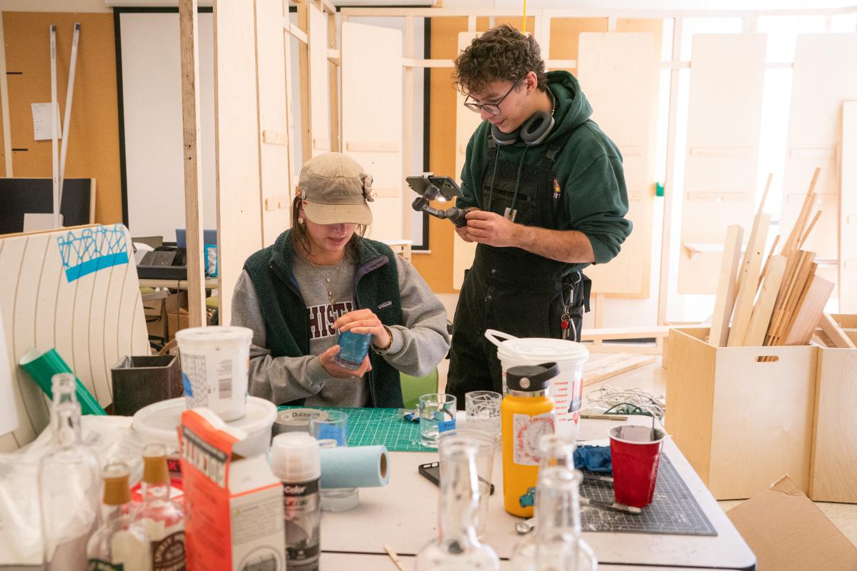 A student works on a project in a shop while another records the work.
