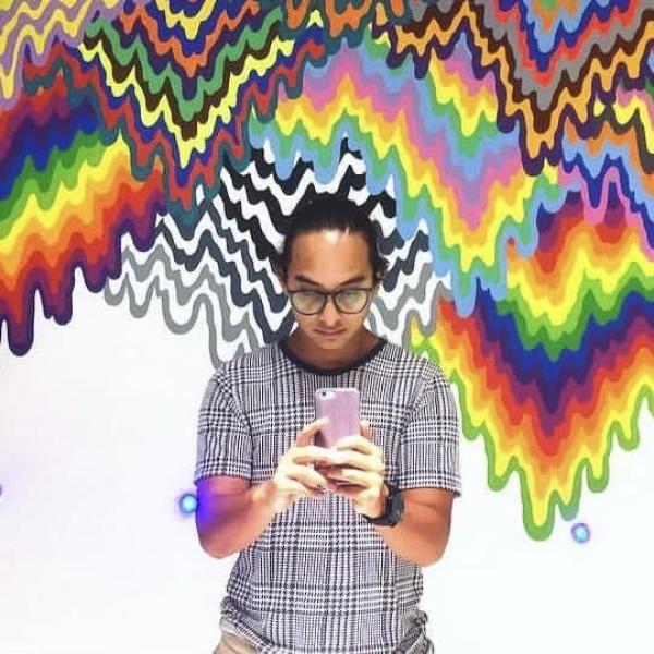 Photo of a man in dark clothes standing in front of a colorfully painted wall.