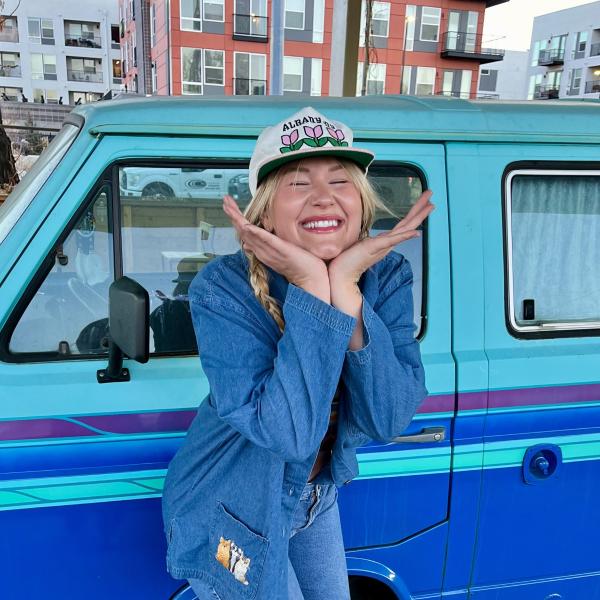 Hannah, a blonde woman with fair skin, smiling and standing in front of a blue car. She is wearing a baseball cap and denim clothing.