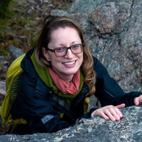 Hilary, who has light colored skin, light brown hair, and glasses. She is smiling while climbing a boulder outdoors.