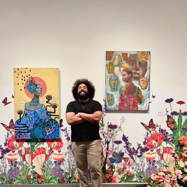 Terrance, who has dark hair and is smiling gleefully while standing between two colorful paintings. There is floral wallpaper behind him. He has an afro, a beard, and is wearing a black T-shirt.