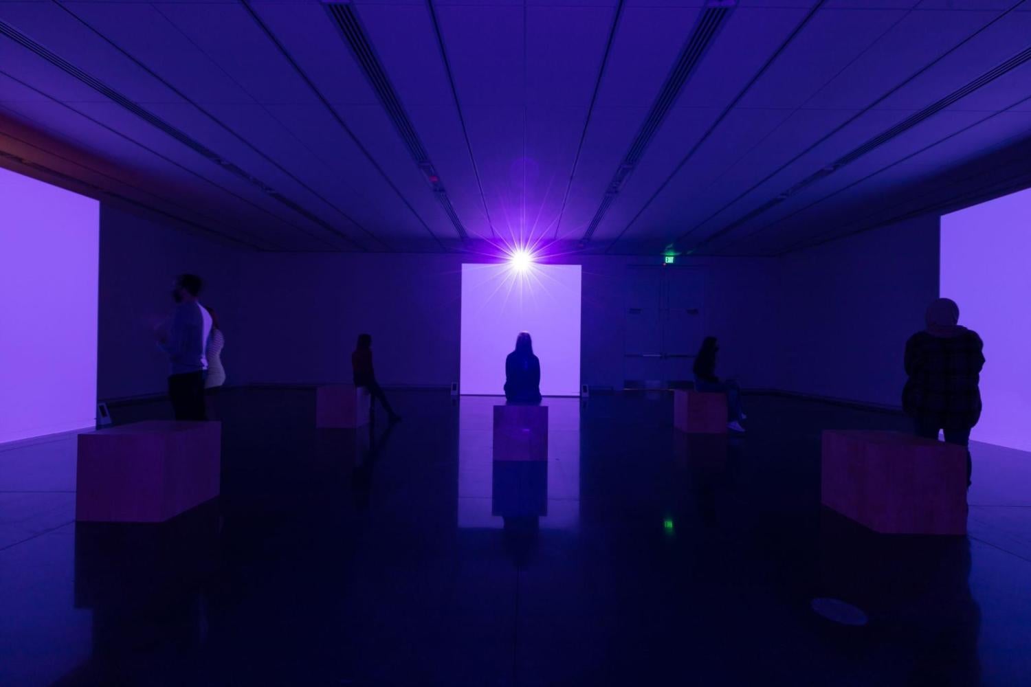 Installation shot of "Lamont Hamilton: To Hear the Earth Before the End of the World." A dark gallery with people standing or sitting. There are three brightly lit purple walls.