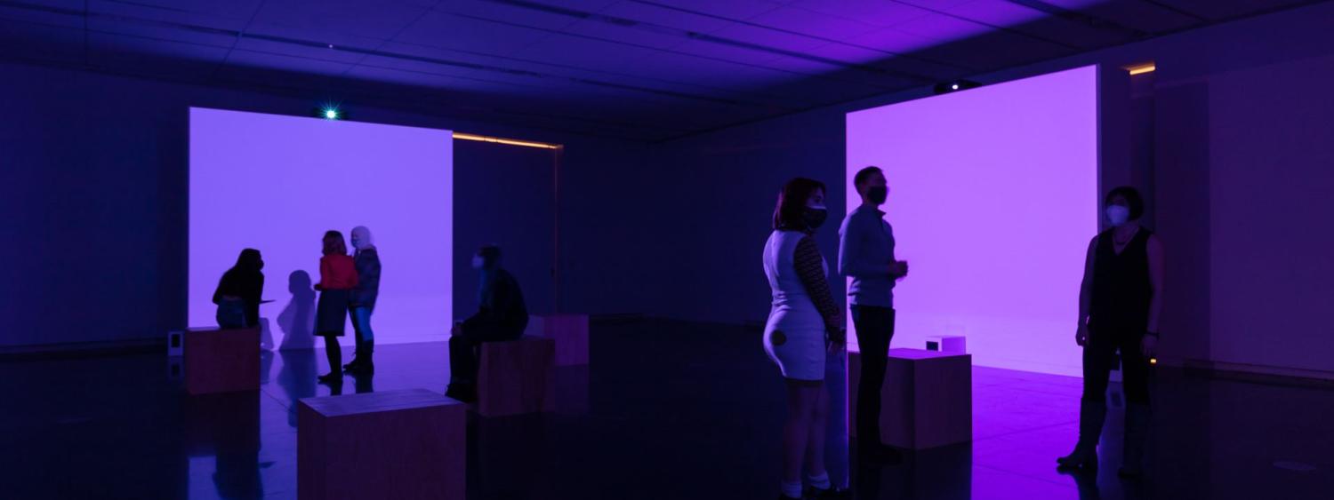 Many people in a dark art gallery at the CU Art Museum with two brightly lit purple walls.