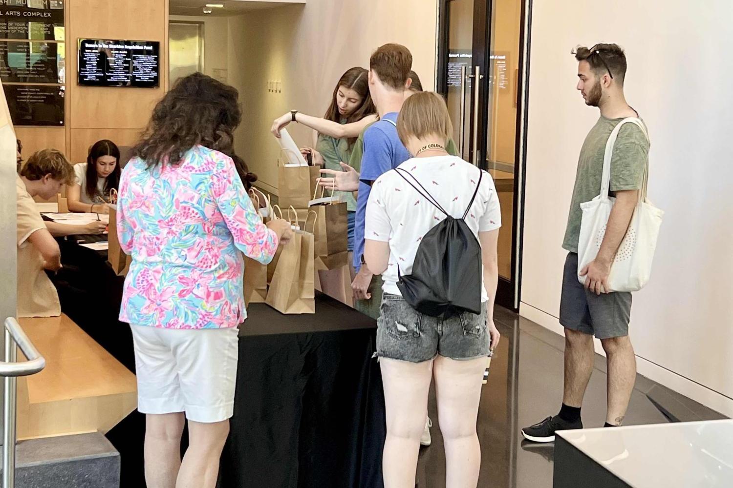 Students in the CU Art Museum lobby taking paper bags filled with art supplies from a table with a black tablecloth on it. In the background, students are seated at another table while coloring.