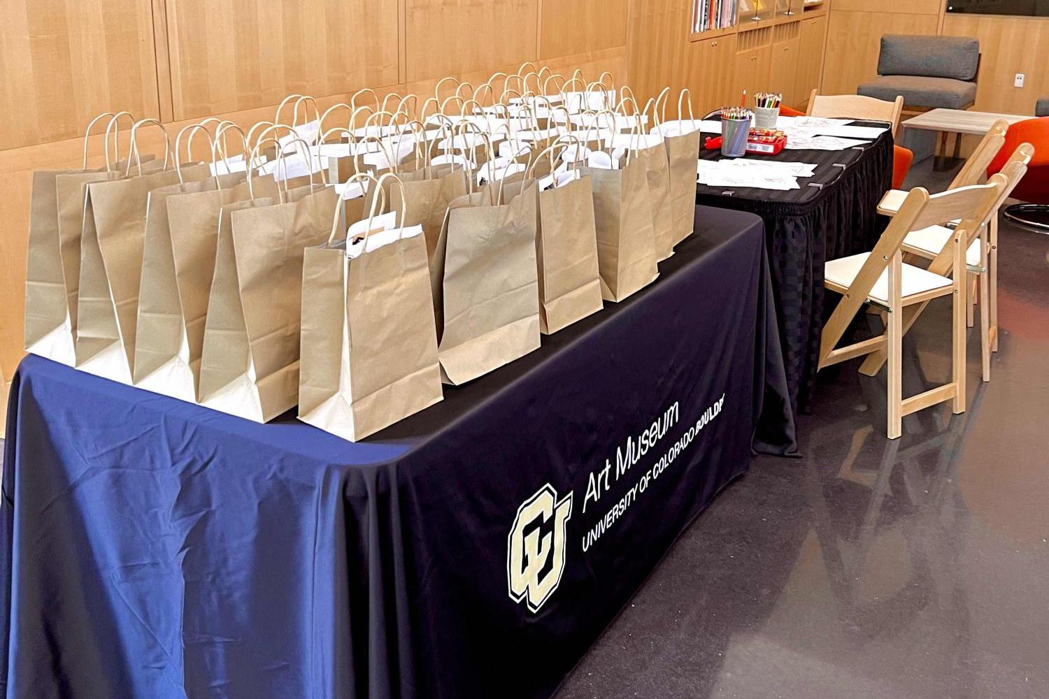 The CU Art Museum lobby with two tables covered in black tablecloths. On the left table are many brown paper bags with art supplies inside, and on the right table there are coloring pages, colored pencils, and crayons.