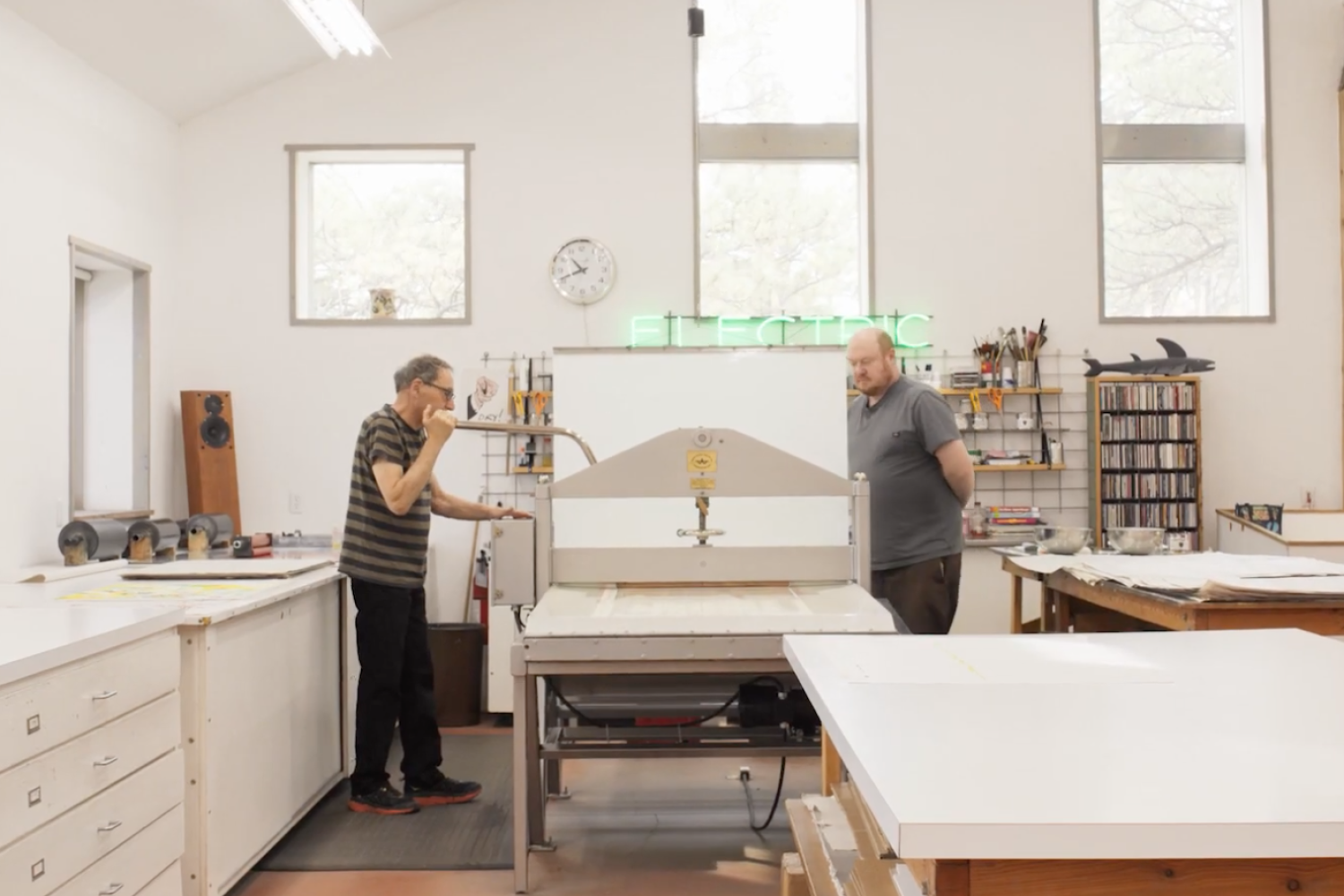 Bud Shark (on the left) and a studio hand (on the right) standing at a lithography printing press in the Shark's Ink print studio. The walls, tables, and cabinets in the room are all white. There is a model shark atop a bookcase next to various brushes, rollers, and other tools. A neon sign in the background reads "electric".