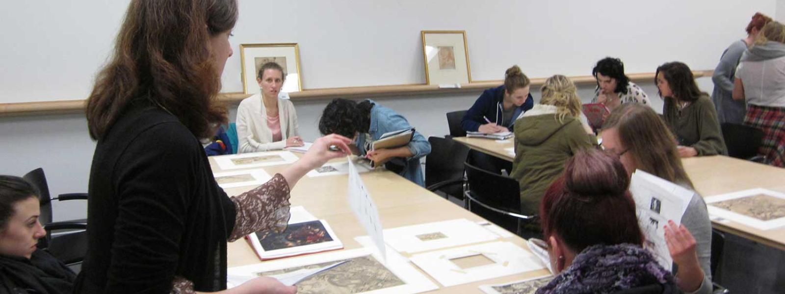 Woman gesturing to prints laying out on a table while speaking to a group of students