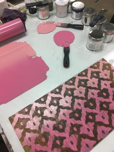 A carved woodblock, roller, ink, and other printmaking tools on a white glass table. Pink ink has is puddled on the table.