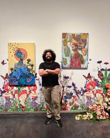 Terrance, who has dark hair and is smiling gleefully while standing between two colorful paintings. There is floral wallpaper behind him. He has an afro, a beard, and is wearing a black T-shirt.