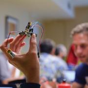 close up of a small device being held between a few fingers, blurred background of people. 