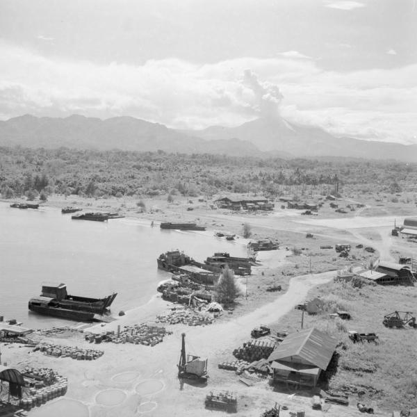 View of Cape Torokina with landing craft in foreground and Mt. Bagana Volcano erupting in background. Torokina Fighter Strip can be seen at the extreme right.