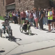 Two students race their vehicles down a sidewalk as their classmates look on