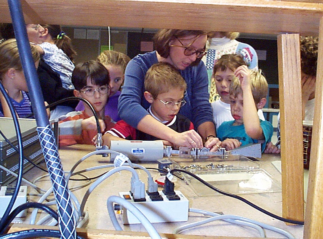 Zoya leans over an elementary-aged child to demonstrate a device to him and his classmates