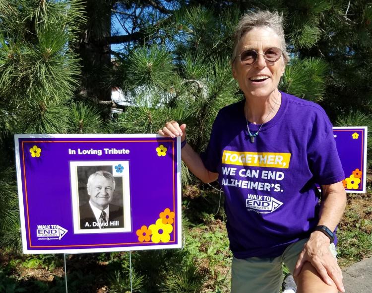 Woman kneels next to poster with a photo of A. David Hill and the words "In Loving Tribute"