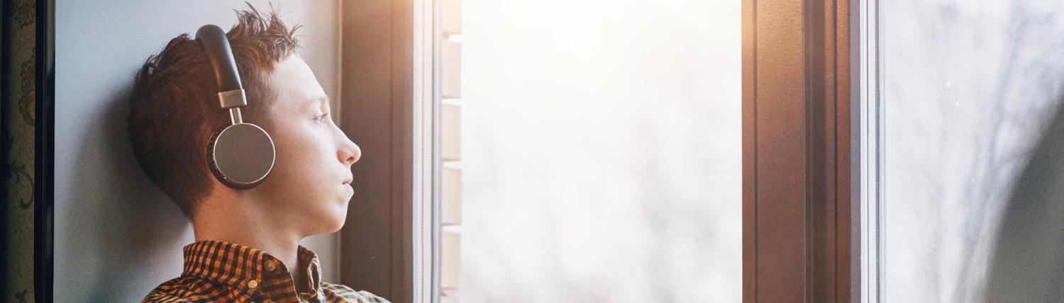 Teenager looking out the window of his room while listening to music with headphones.