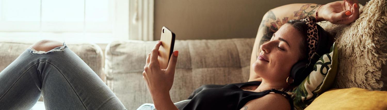 Girl laying on couch wearing headphones in a relaxed position looking at her phone. 