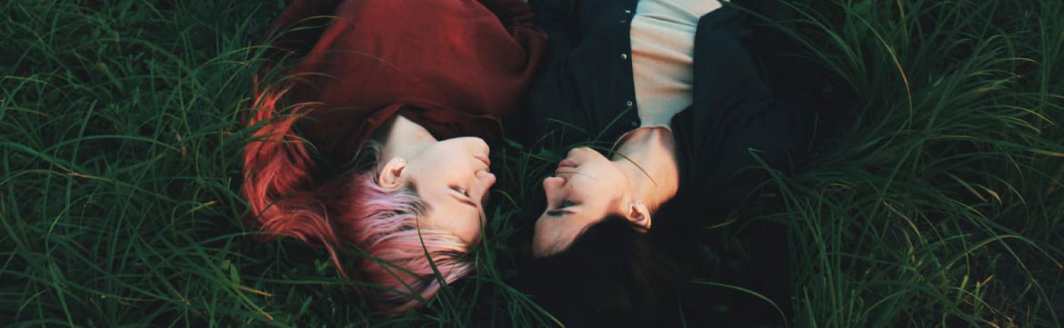 Photo of two people laying together in the grass as they look at each other.
