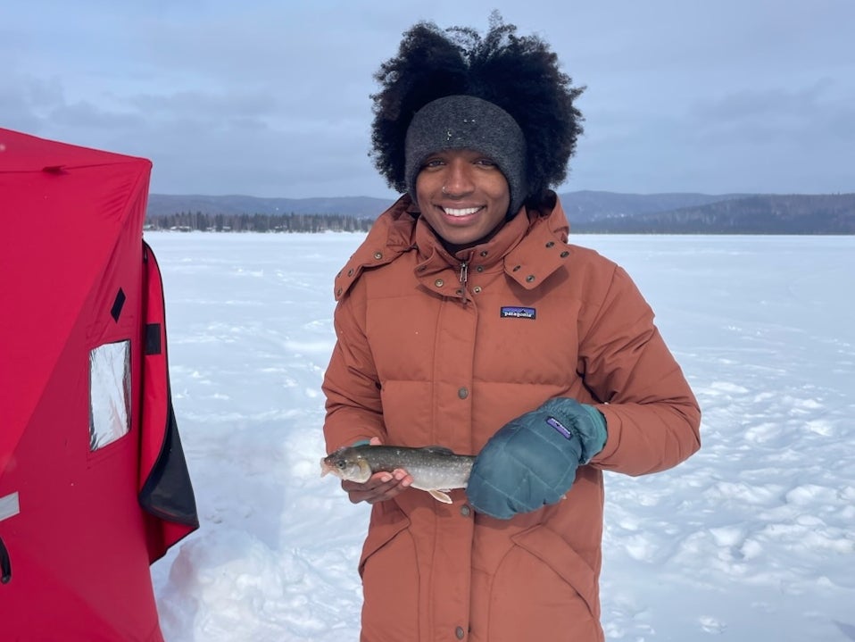 Peyton Thomas in a long puffy jacket holds a recently caught fish on a frozen lake with a red tent beside her.