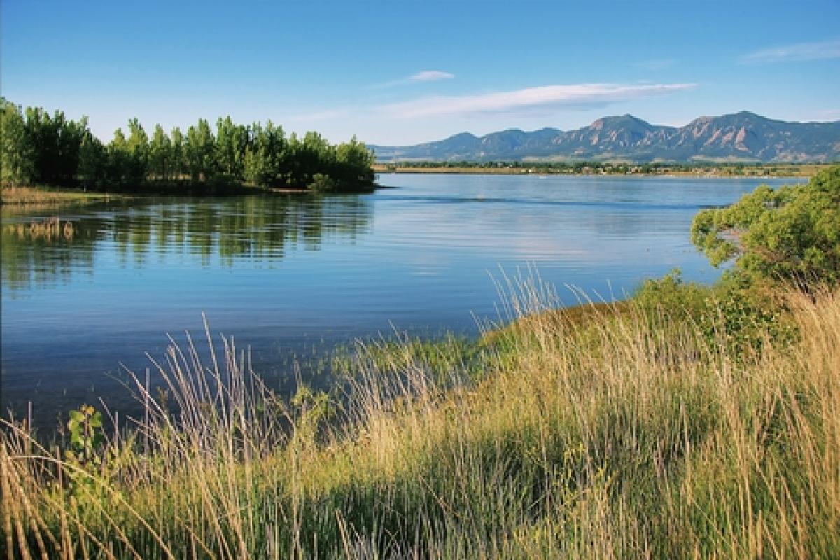Grass lined banks and blue waters of the Boulder Reservoir on a summer morning, with the jagged flatirons far in the background. Photo by Richard Saxon (Flickr)