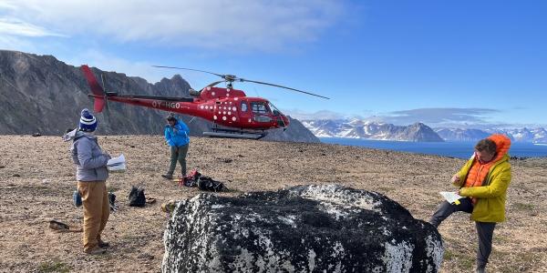 Three researchers gather around a very large boulder on a flat surface high above the ocean.  A red helicopter is behind them.  In the distance are rugged mountains and fjords.