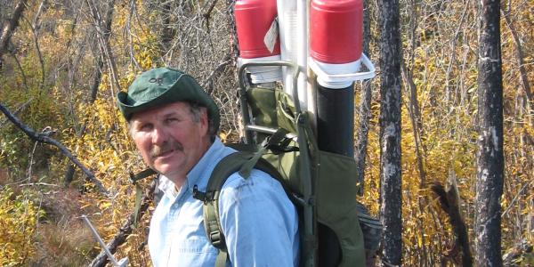 George Aiken gears up for fieldwork, wearing a backpack with long tubes and instruments, and surrounded by other equipment