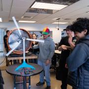 A wind team member in a propeller beanie demonstrates a tabletop turbine model during at K-12 outreach event