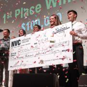 A diverse team of students stands on stage with their giant prize-winning check, the air is full of confetti, their joy is infectious